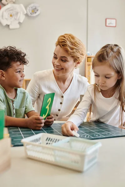 Happy african american boy holding carton with number near girl and teacher in Montessori school — Stock Photo