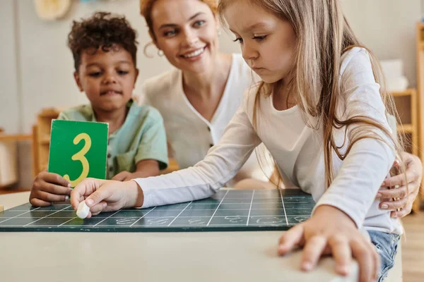 Girl writing on chalkboard near african american boy with number and teacher, Montessori school — Stock Photo