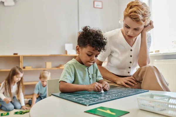 Teacher talking to african american boy writing on chalkboard, counting, learning through play — Stock Photo