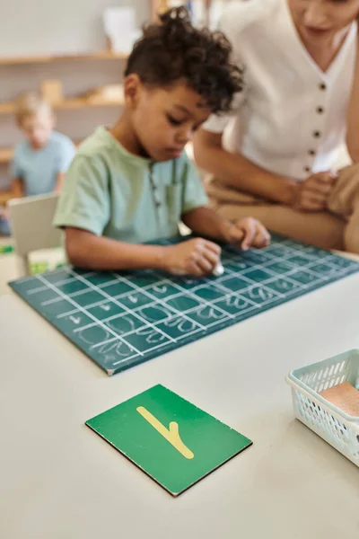 Teacher observing african american boy writing on chalkboard, counting, learning through play — Stock Photo