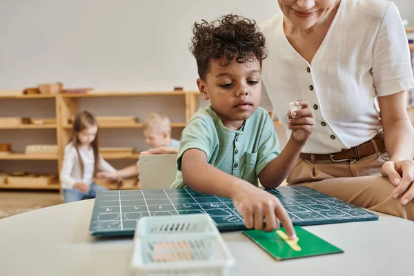 Cheerful teacher observing african boy pointing at number near chalkboard, learning through play — Stock Photo