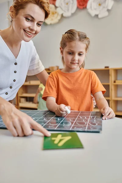 Happy teacher pointing at number near smart girl in Montessori school, counting, writing, chalkboard — Stock Photo