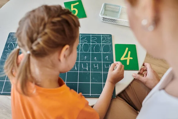 Top view of clever girl writing on chalkboard, learning how to count near teacher, Montessori school — Stock Photo