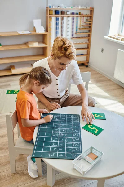 Nettes Mädchen, das auf Kreidetafel neben Zahlen schreibt, spielerisch lernt, Lehrerin, Montessori-Schule — Stockfoto