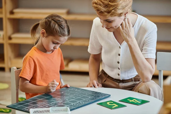 Cute girl looking at chalkboard near numbers, aprender através do jogo, professor, escola Montessori — Fotografia de Stock
