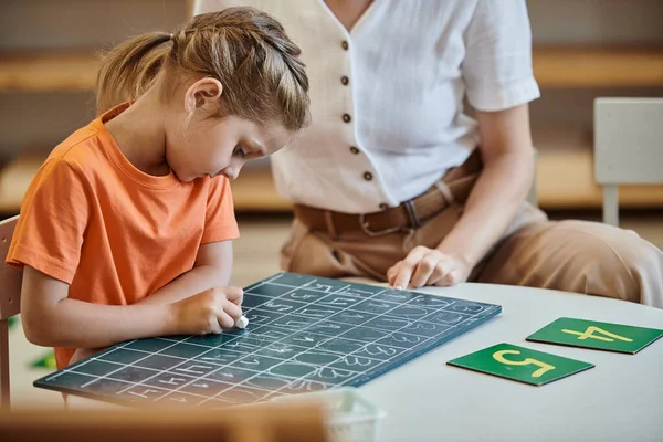 Nettes Mädchen, das auf Kreidetafel neben Zahlen schreibt, spielerisches Lernen, Lehrer, Montessori-Konzept — Stockfoto