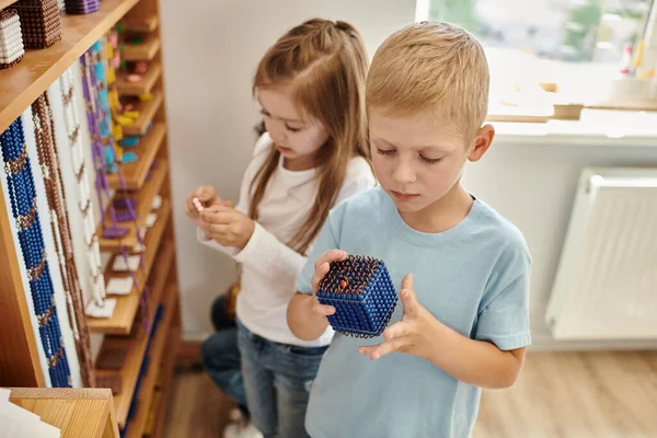 Menino curioso segurando Montessori pérola grânulos material, aprender matemática, menina no fundo borrado — Fotografia de Stock