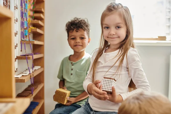 Fille gaie tenant cube avec des perles près du garçon afro-américain, diversité, école Montessori — Photo de stock