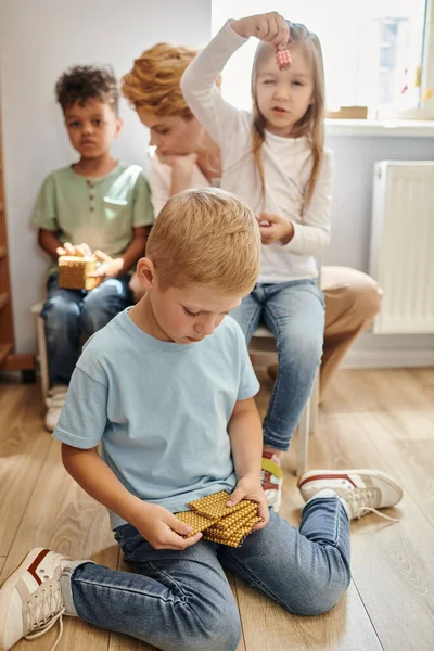 Boy sitting on floor near interracial kids and female teacher, Montessori material, early education — Stock Photo
