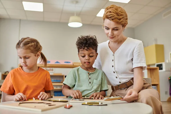 Professeur utilisant matériel didactique montessori tout en jouant avec interracial garçon et fille, divers — Photo de stock