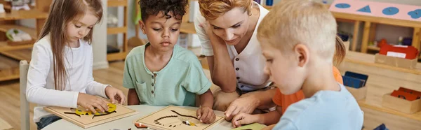 Вчитель використовує дидактичний матеріал під час гри з мультиетнічними дітьми в школі Монтессорі, банер — стокове фото