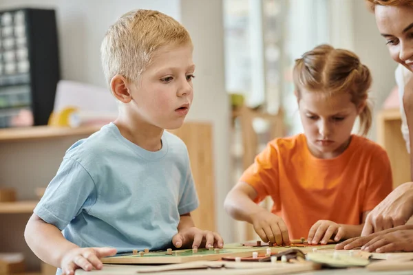 Boy sitting near teacher and didactic materials during lesson in montessori school — Stock Photo