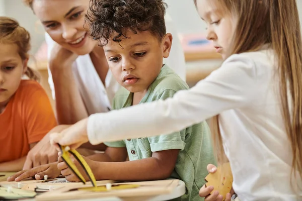 Niños multiétnicos jugando juntos cerca de profesor borroso en la escuela montessori - foto de stock