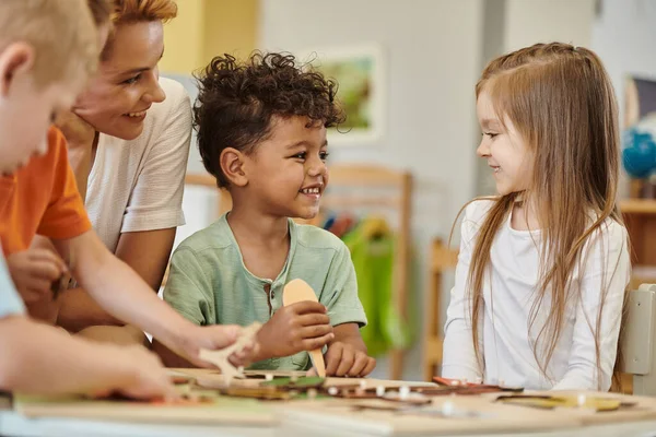 Enfants multiethniques joyeux jouant près de l'enseignant pendant les cours à l'école montessori — Photo de stock