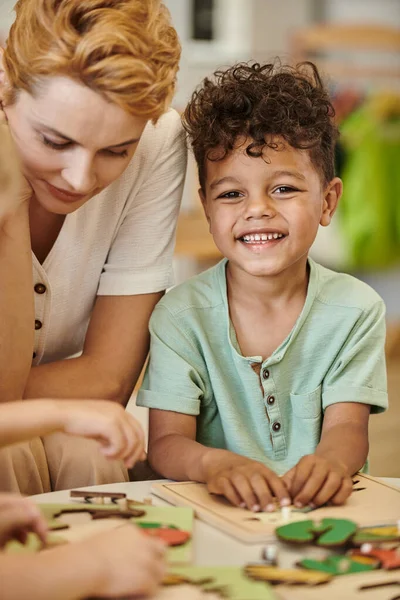 Cheerful african american boy playing near teacher and looking at camera in montessori school — Stock Photo
