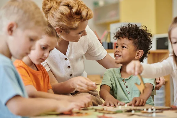 Учитель разговаривает с улыбающимся африканским американским мальчиком, пока дети играют в школе Монтессори — стоковое фото