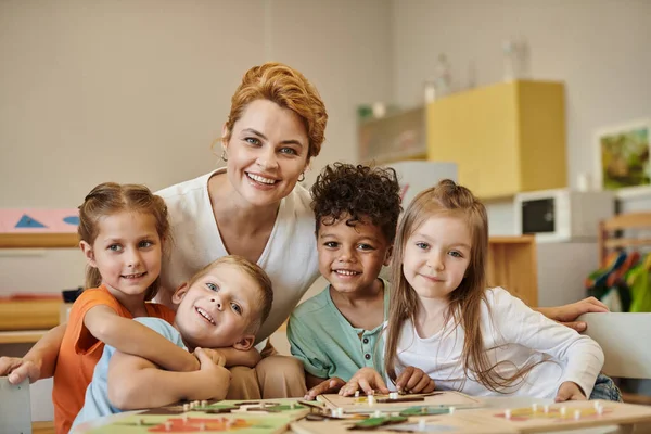 Lächelnder Lehrer und multiethnische Kinder blicken während des Unterrichts in der Montessori-Schule in die Kamera — Stockfoto