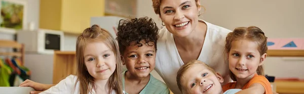 Profesor positivo y niños interracial mirando a la cámara en la escuela montessori, pancarta - foto de stock