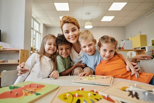 Enseignant souriant étreignant les enfants multiethniques près du matériel didactique sur la table à l'école montessori — Photo de stock