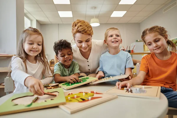 Alegre profesor sentado cerca de multiétnicos niños jugando con material didáctico en montessori clase - foto de stock