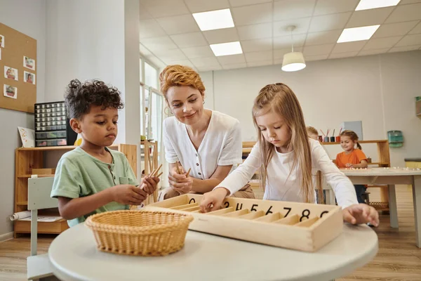 Lächelnder Lehrer neben interrassischen Kindern mit Holzstäben während des Unterrichts in der Montessori-Schule — Stockfoto