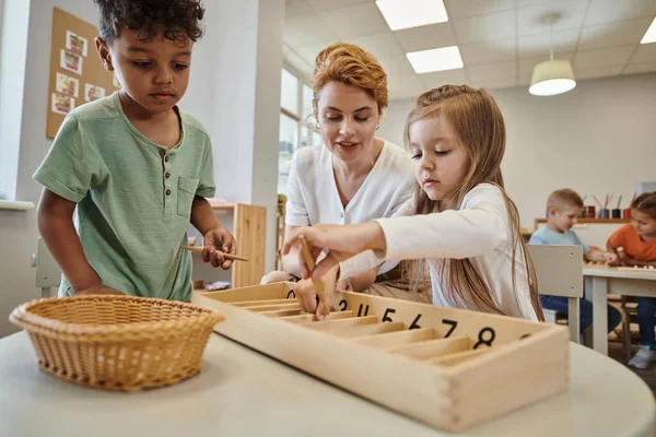 Lehrerin im Gespräch mit interrassischen Kindern, die in Montessori-Schule mit Holzstäben spielen — Stockfoto