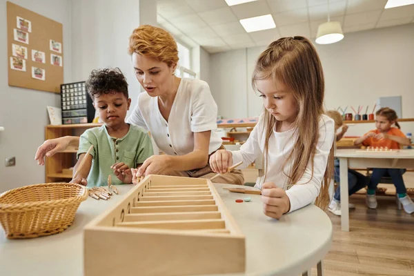 Teacher talking to multiethnic kids while playing with wooden sticks in montessori school — Stock Photo