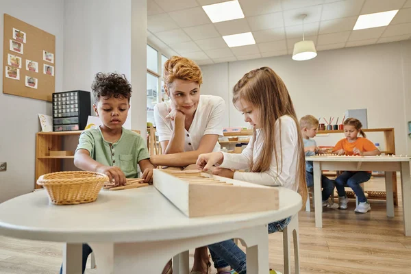 Teacher sitting near multiethnic kids playing with wooden sticks during lesson in montessori school — Stock Photo