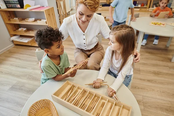 Vista de ángulo alto del profesor sonriente abrazando a niños multiétnicos jugando con palos en la escuela montessori - foto de stock
