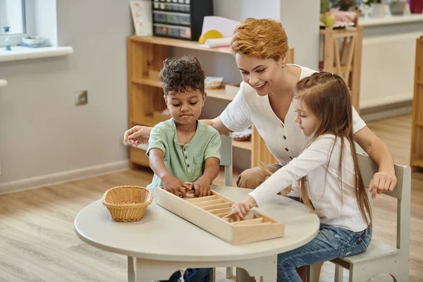Insegnante felice parlando con i bambini interrazziali che giocano con materiali didattici in legno in classe montessori — Foto stock