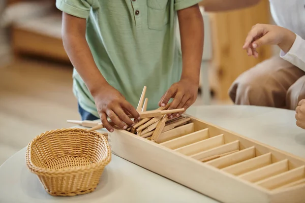 Vista recortada de chico afroamericano sosteniendo palos de madera cerca de chica en la escuela montessori - foto de stock