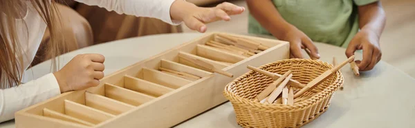 Vista recortada de niños interracial jugando con palos de madera en la escuela montessori, pancarta - foto de stock