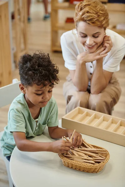 Professeur positif assis près d'un enfant afro-américain jouant avec des bâtons en bois à l'école montessori — Photo de stock