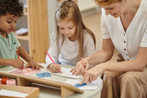 Мультиэтнические дети рисуют карандашами и разговаривают рядом с улыбающимся учителем в школе Монтессори — стоковое фото