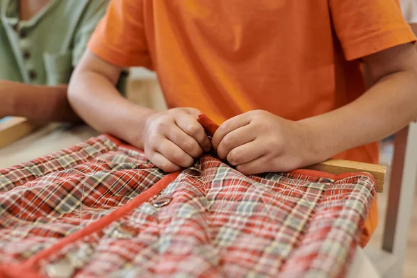 Vista ritagliata di bambino abbottonatura panno su telaio vicino amico durante la lezione a scuola montessori — Foto stock