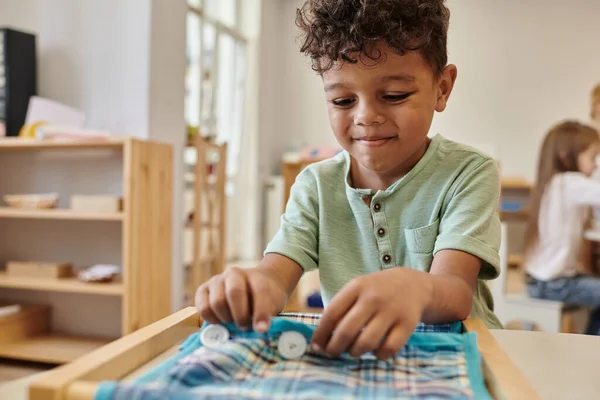 Smiling african american boy playing with cloth and buttons in montessori school — Stock Photo