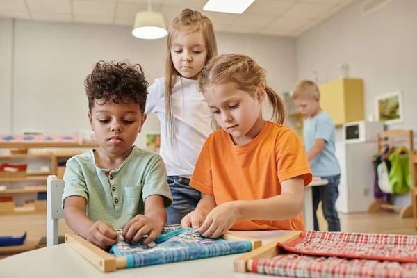 Межрасовые дети играют с тканью и пуговицами на рамке в классе Монтессори школы — стоковое фото