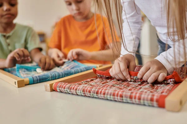 Обрезанный вид ребенка, играющего с тканью и пуговицами рядом с многонациональными друзьями в школе Монтессори — стоковое фото