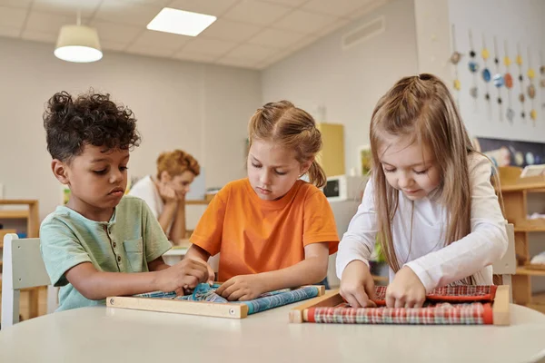 Multiethnic kids playing with cloth and buttons on table in montessori school — Stock Photo
