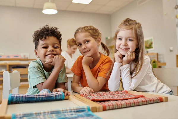 Niños alegres y multiétnicos mirando la cámara cerca de la mesa en la clase de la escuela montessori - foto de stock