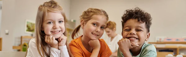 Bambini sorridenti e interrazziale guardando la fotocamera in classe offuscata nella scuola montessori, banner — Foto stock