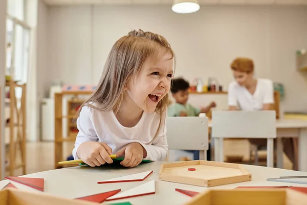 Niño alegre mirando lejos cerca de materiales didácticos en la clase de la escuela montessori - foto de stock