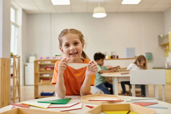 Enfant positif tenant triangle en bois et regardant la caméra dans l'école montessori — Photo de stock