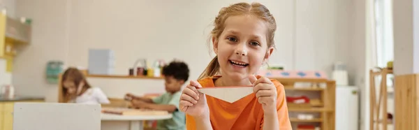 Cheerful girl looking at camera and holding triangle in montessori school, banner — Stock Photo