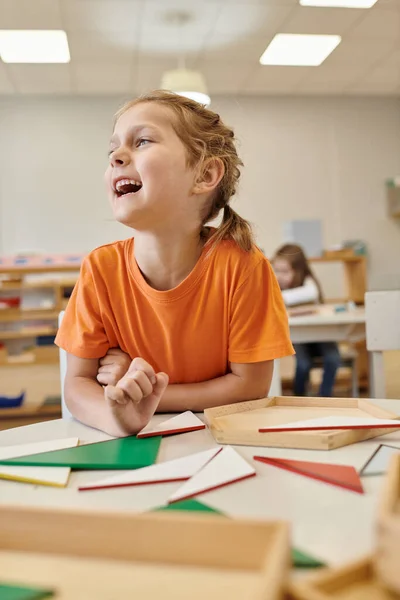 Niño feliz mirando lejos cerca de materiales didácticos de madera en la clase de la escuela montessori - foto de stock
