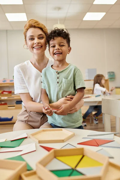 Усміхнений вчитель обіймає афроамериканську дитину біля дидактичних матеріалів у школі Монтессорі — стокове фото