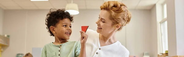 Heureux garçon afro-américain tenant triangle près de l'enseignant pendant les cours à l'école montessori, bannière — Photo de stock