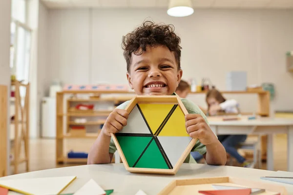 Menino afro-americano positivo segurando materiais didáticos de madeira na escola montessori turva — Fotografia de Stock