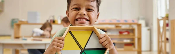 Menino americano africano feliz segurando jogo de madeira na escola montessori turva, bandeira — Fotografia de Stock