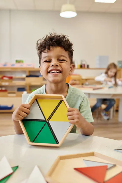 Heureux garçon afro-américain tenant jeu en bois près de la table en classe à l'école montessori — Photo de stock